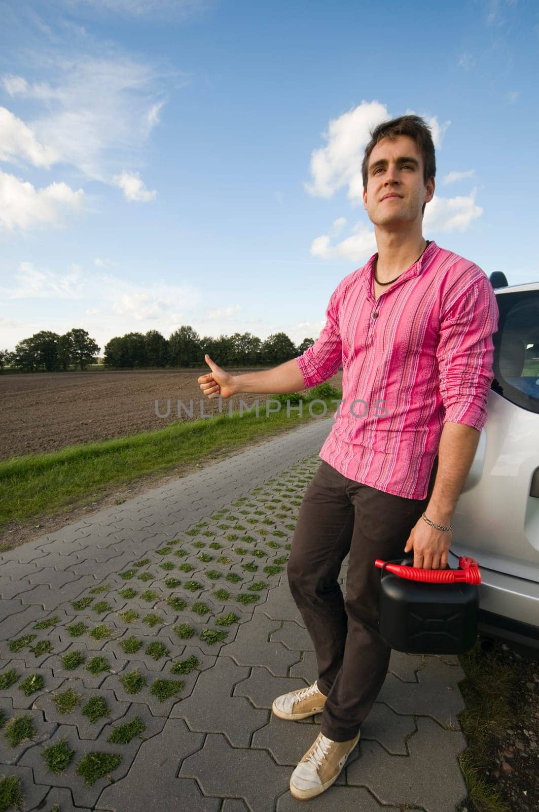Young man with black reserve canister on a lonely country road leans helplessly against a car and waits for help