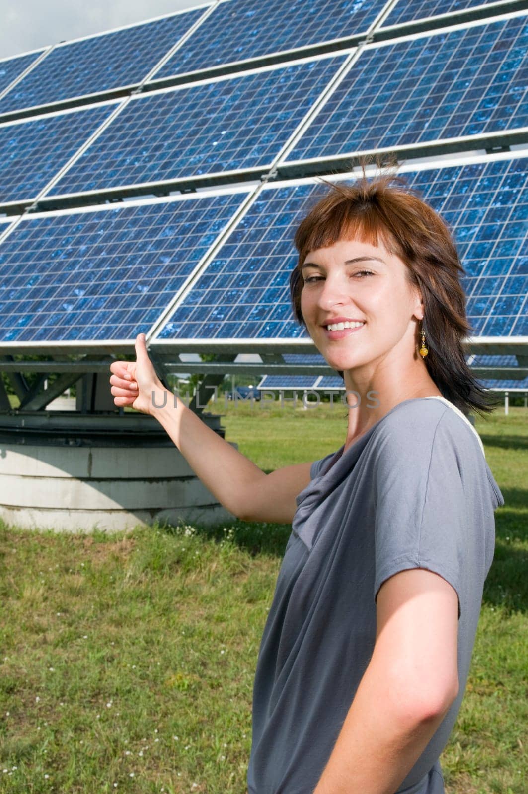 Waist up of a young, brunette woman in a casual leisure dress on a green meadow in summer, presenting and pointing to a photovoltaic system behind her