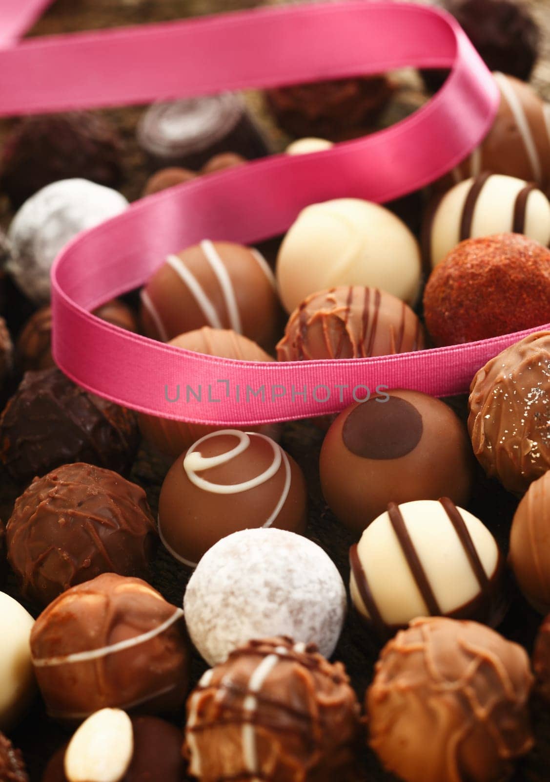 A decorative pink ribbon lies twirled over handmade spherical chocolates with different patterns in a gift and celebration concept