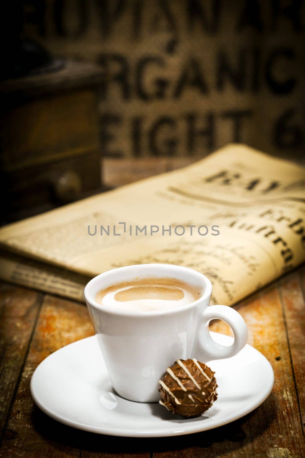 Cup of rich milky coffee with a chocolate bonbon on the saucer and a folded newspaper on the table behind in a coffee break concept