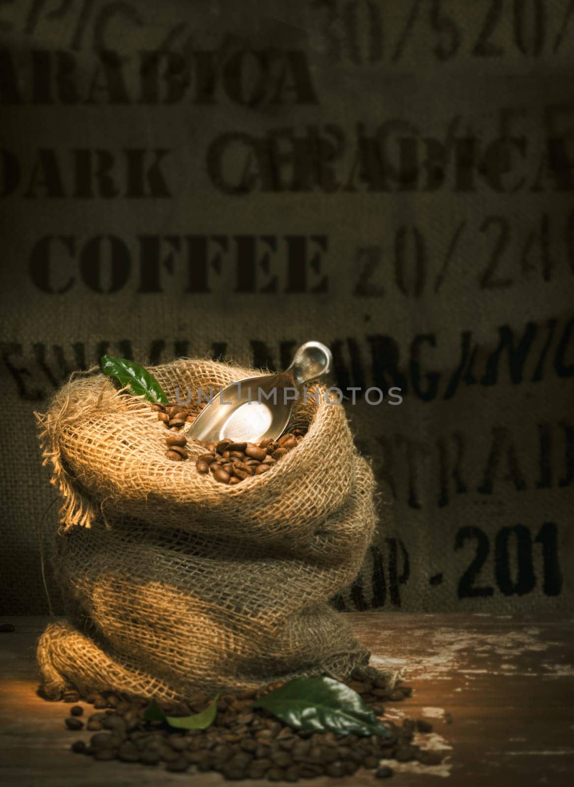 Small rustic hessian bag filled to overflowing with fresh roasted coffee beans and topped with a small trowel and green leaves on a dark background with copyspace