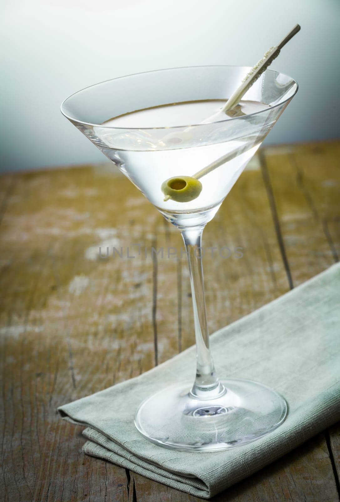 Clear alcoholic cocktail with an olive in a cocktail glass standing on a napkin over rustic wooden boards