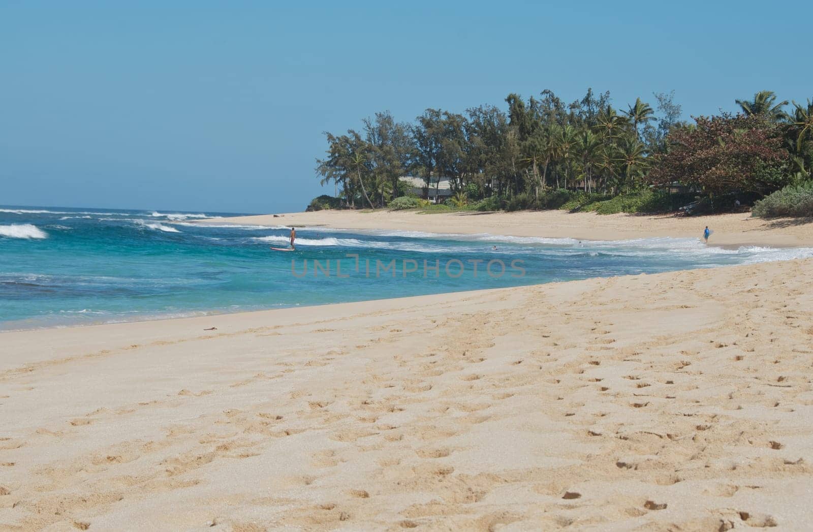 Idyllic tropical sandy beach and bay with a lone surfer in the distance amongst the breaking waves.