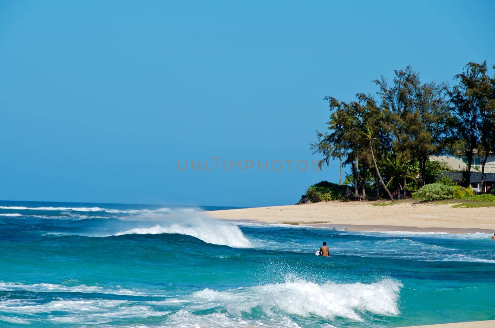 Lone surfer amongst breaking waves and white surf in an idyllic turquoise blue tropical bay with sandy beach