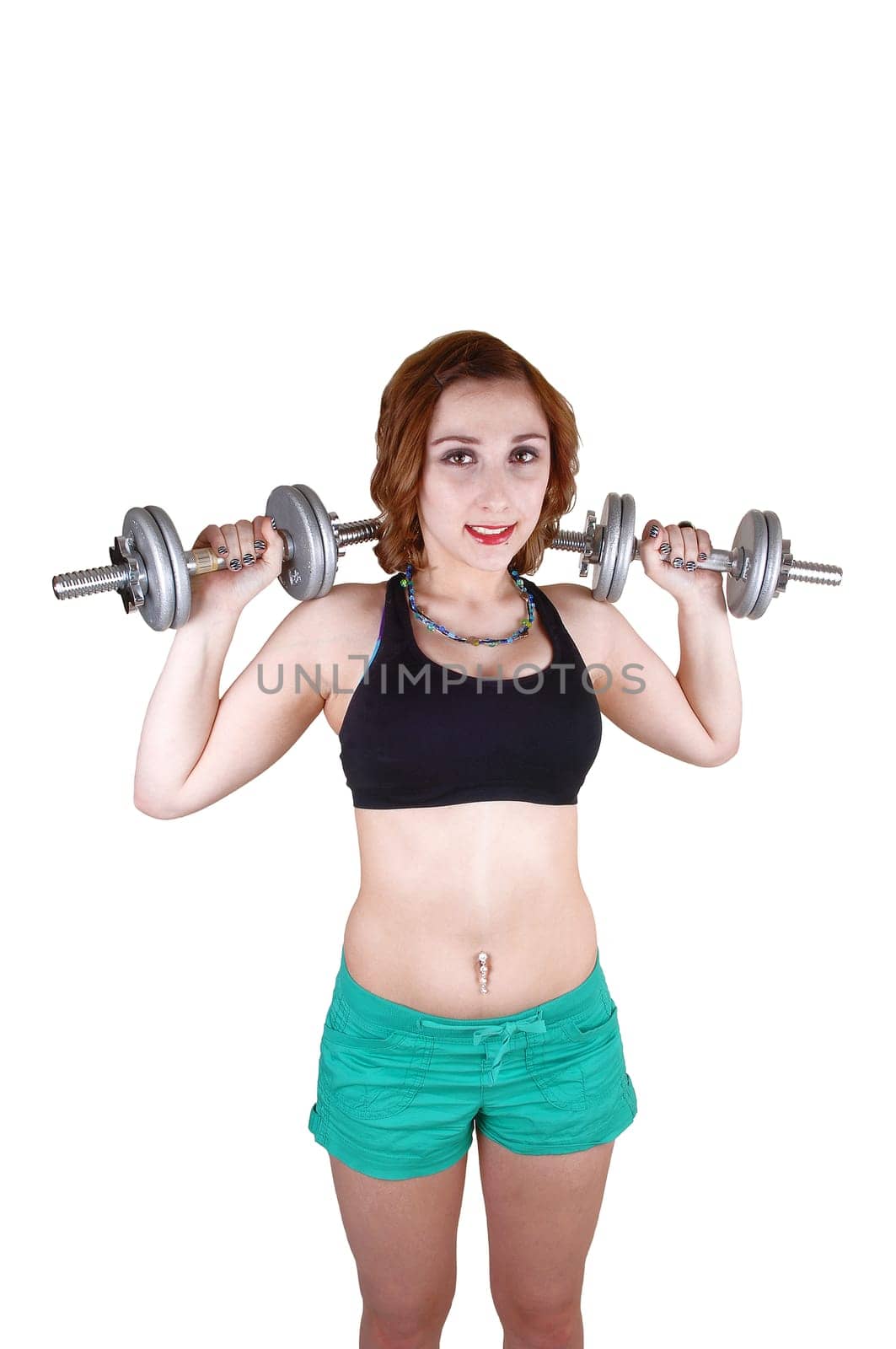 A teenage girl standing for white background in green shorts and a black sports bra, lifting up two drumbells for her exercise.