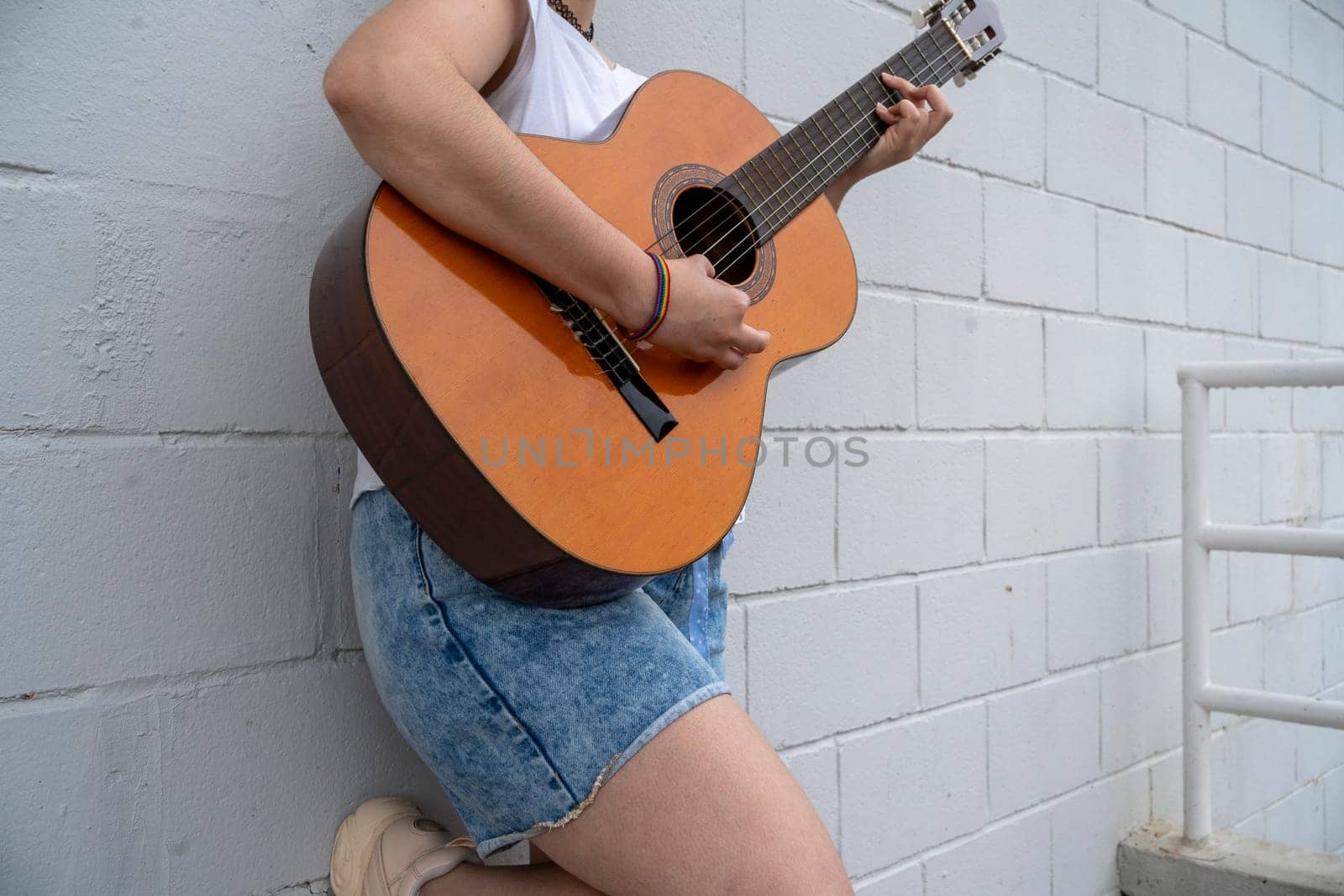 Crop anonymous female guitarist with rainbow bracelet playing music on acoustic guitar while standing near metal fence on street in public place