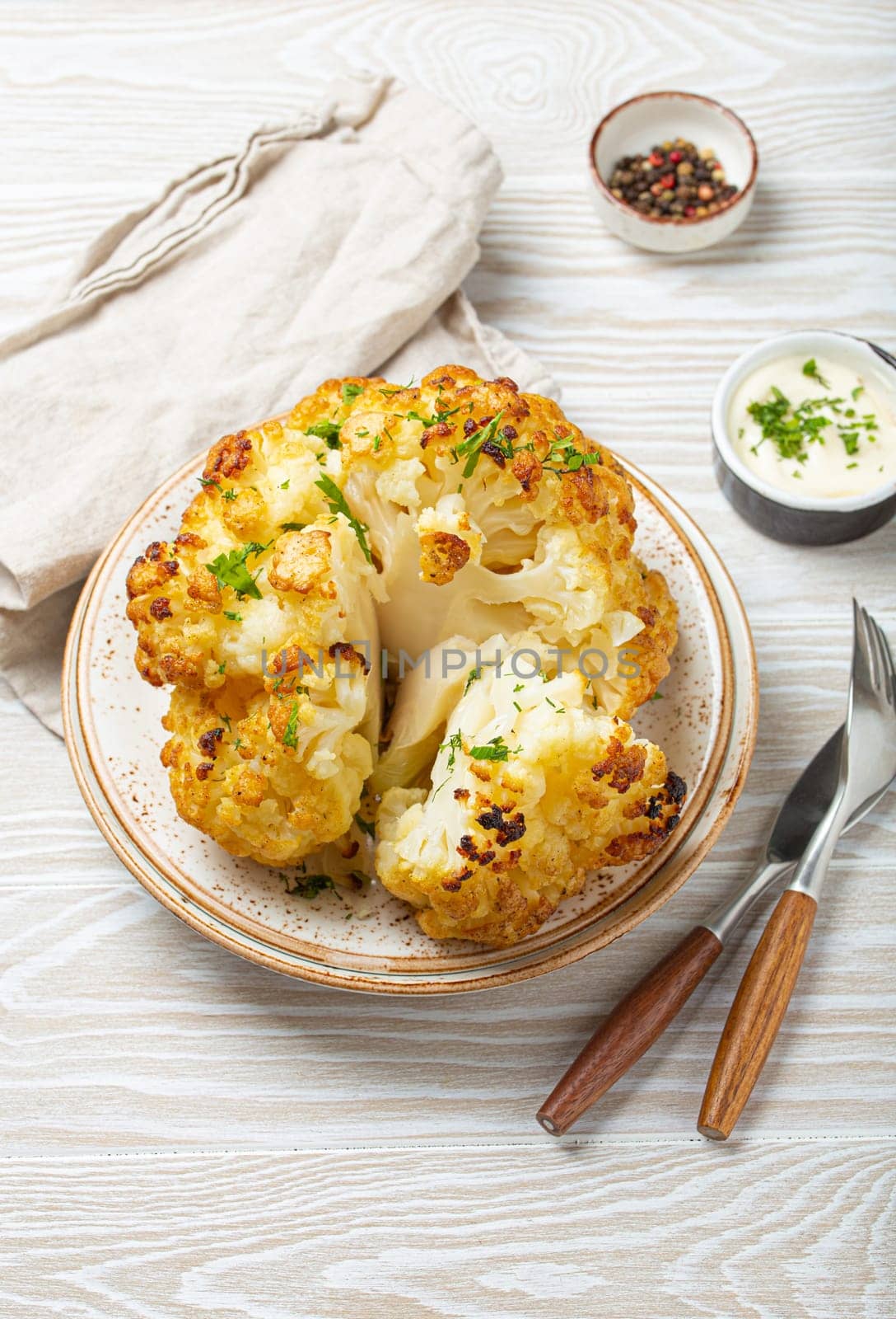 Whole roasted head of cauliflower with olive oil on plate on white wooden rustic table top view with cutlery. Vegetarian dish. by its_al_dente