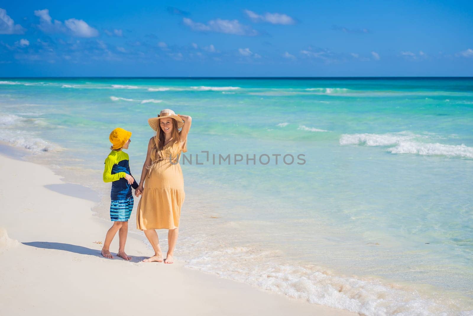 A radiant pregnant mother and her excited son share a tender moment on a serene, snow-white beach, celebrating family love amidst nature's beauty.