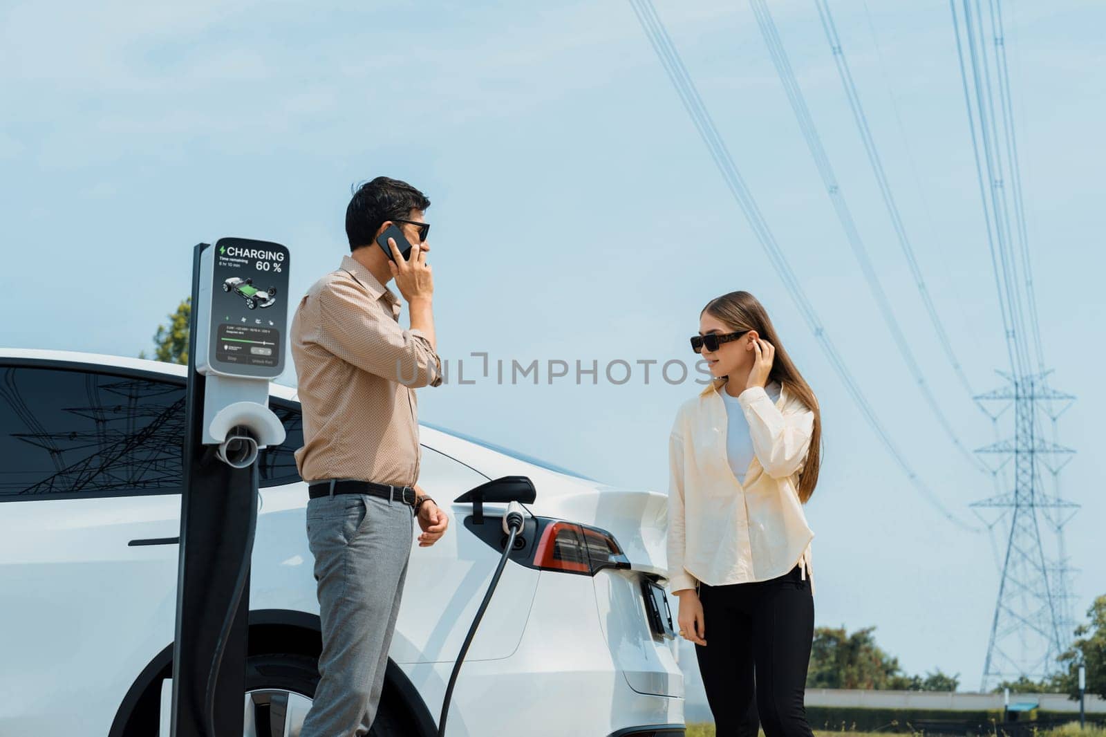 Couple pay for electricity with smartphone while recharge EV car battery at charging station connected to power grid tower electrical as electrical industry for eco friendly car utilization.Expedient
