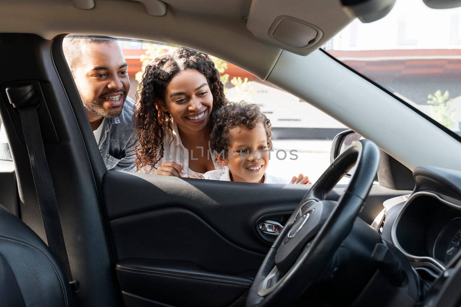 Happy african american family choosing new car at dealership center parking spot. Excited black mom, dad and preteen child son looking inside new automobile and smiling. Auto buying, transportation