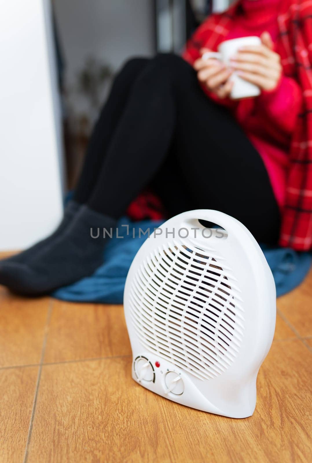 A woman in a plaid and a warm sweater warms herself at home near an electric fan heater, holds a cup of hot tea in her hands, close-up