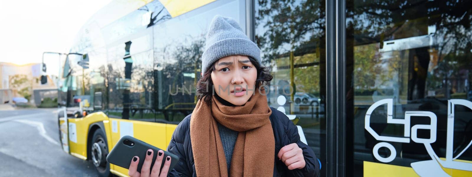 Portrait of confused asian girl, standing on bus stop, holding mobile phone, looking shocked and upset, disappointed by public transport delay by Benzoix