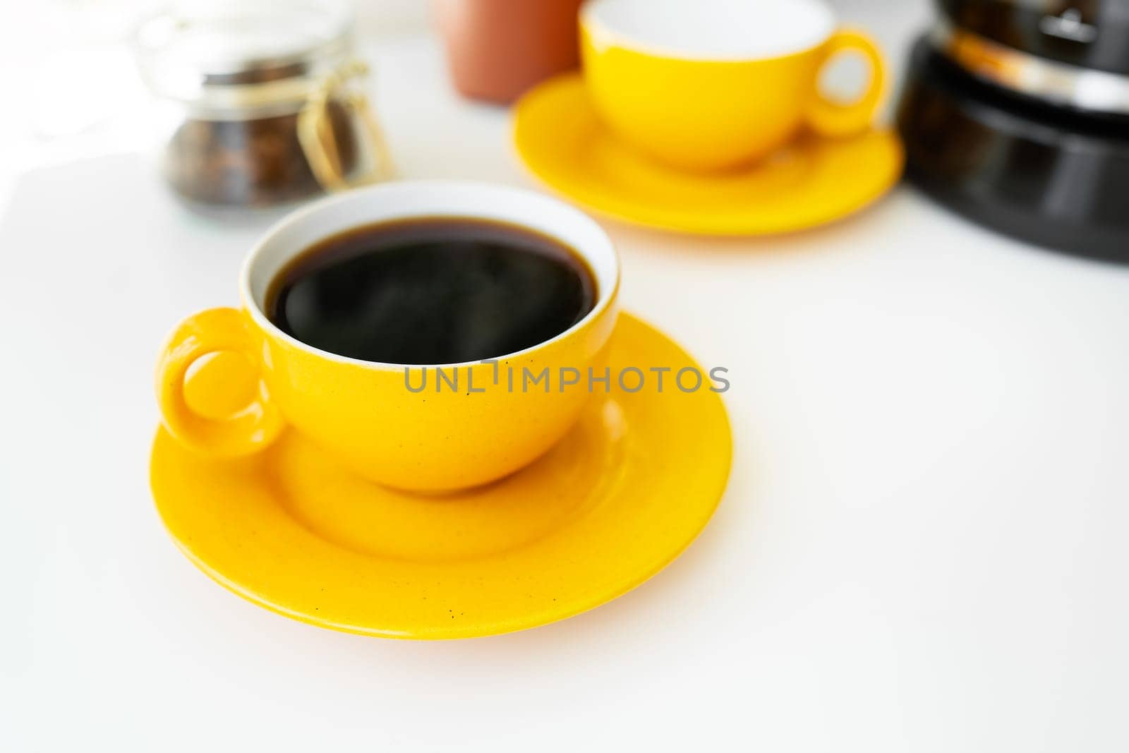 Hot aromatic black coffee in a bright yellow cup and saucer stands on a white table against the background of a coffee maker and a jar of coffee beans. Place for an inscription