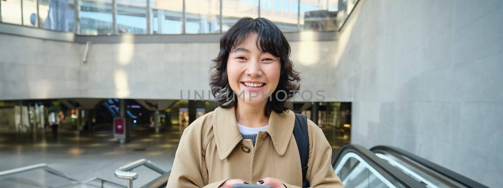 People in city. Young korean woman travels around city, goes up the escalator, uses her mobile phone and smiles.