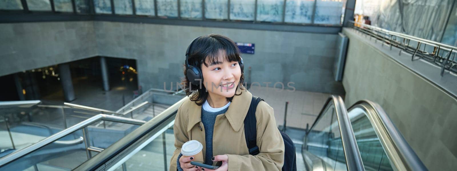 Portrait of smiling korean girl drinks coffee to go, goes up an escalator, holds smartphone, visits new city, arrives at train station, listens music in headphones.