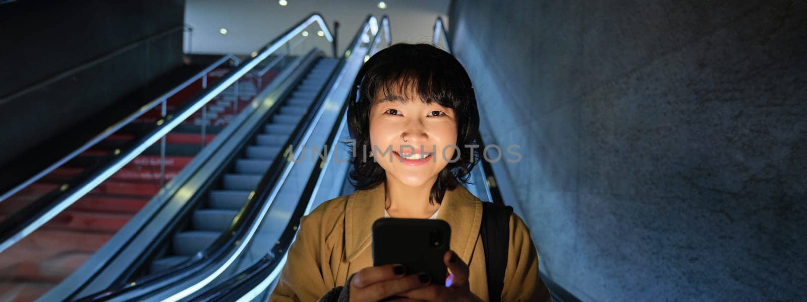 Portrait of girl student in headphones, listens music, commutes, goes down escalator, looks at smartphone with pleased smile, happy face by Benzoix