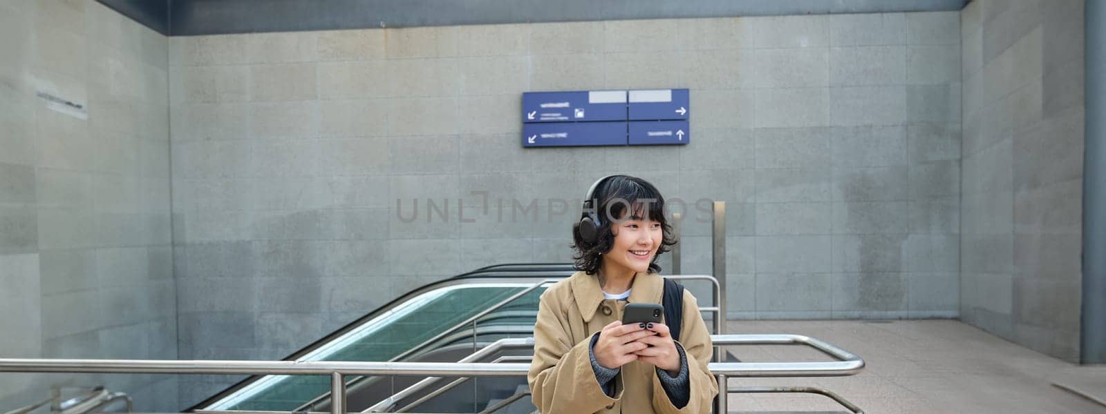 Portrait of stylish asian hipster girl, listens music in headphones, looks at her phone, tourist looking at map for a way to sightseeing, drinking coffee to go.