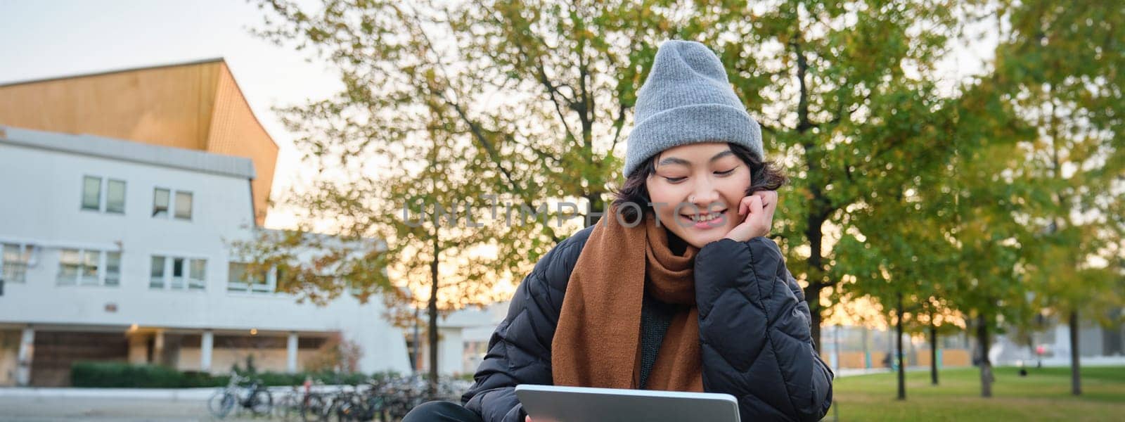 Portrait of asian girl relaxing in park, watching videos or reading on digital tablet, sitting on bench in hat and scarf on chilly day, enjoing outdoors.