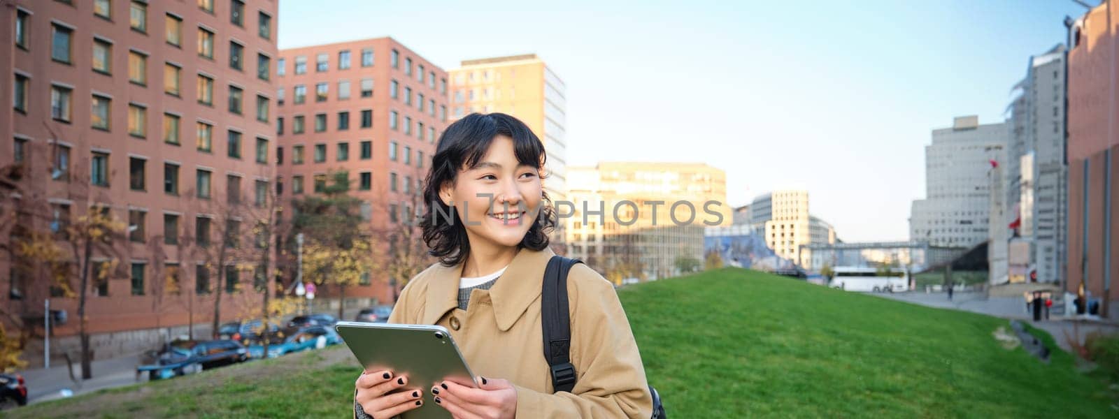 Happy young brunette girl, asian woman walks around city with tablet, goes to university with her digital gadget and backpack.