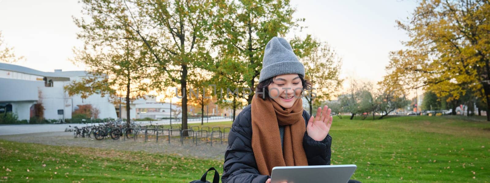 Portrait of young asian girl sits in warm clothes in park, waves hand at tablet, video chats outdoors, says hello while on call by Benzoix