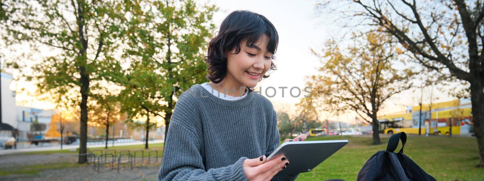 Portrait of young smiling korean girl, graphic designer, artist drawing on digital tablet with a pen tool, sitting in park on fresh air and scatching, taking notes.