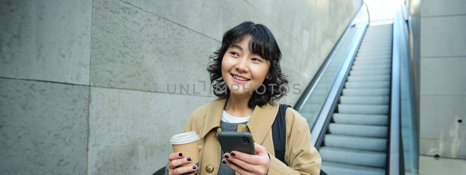 Beautiful brunette girl drinks coffee to go, goes down escalator and smiles, holds smartphone, uses mobile app. Copy space