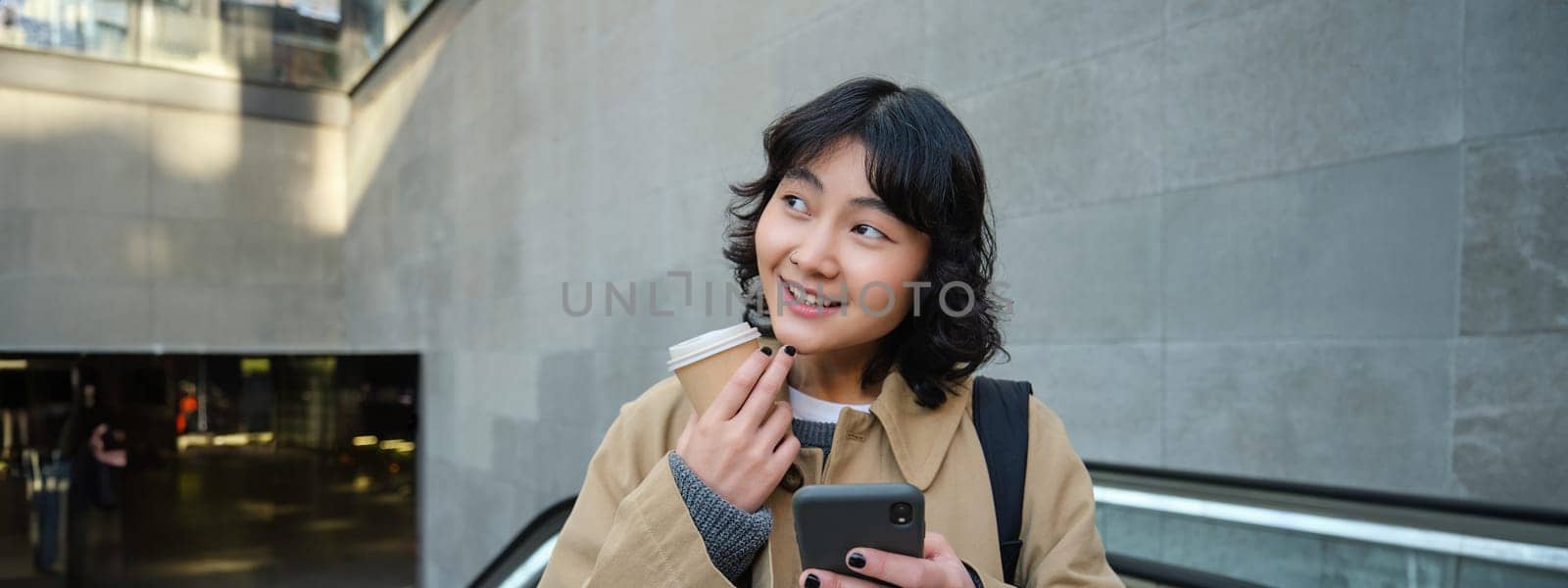 Beautiful happy asian girl, drinks coffee to go, using mobile phone while standing on escalator, walking in city centre and smiling.