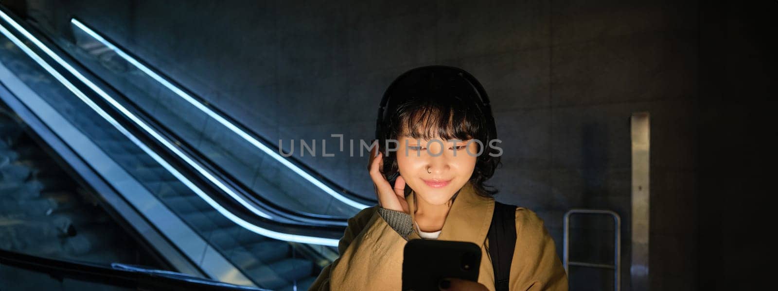 Portrait of beautiful korean girl in headphones, stands near escalator, travels, commutes home on public transport, holds smartphone, poses with mobile phone by Benzoix