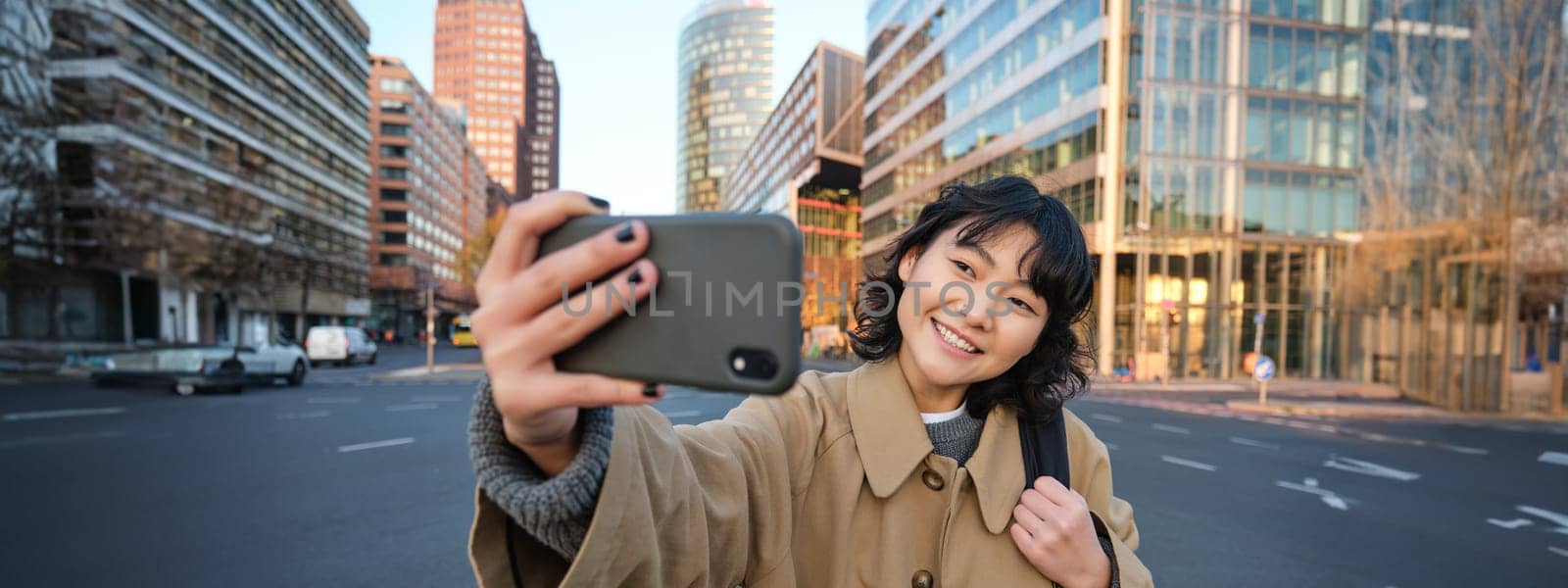 Portrait of young asian woman taking selfie in front of building in city centre, tourist takes photos while sightseeing, smiling at smartphone camera by Benzoix