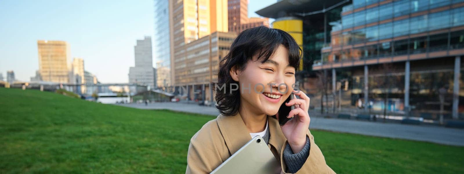 Stylish smiling girl, university student in trench coat, holds tablet, talks on mobile phone, has conversation over telephone and looks relaxed, stands on street by Benzoix