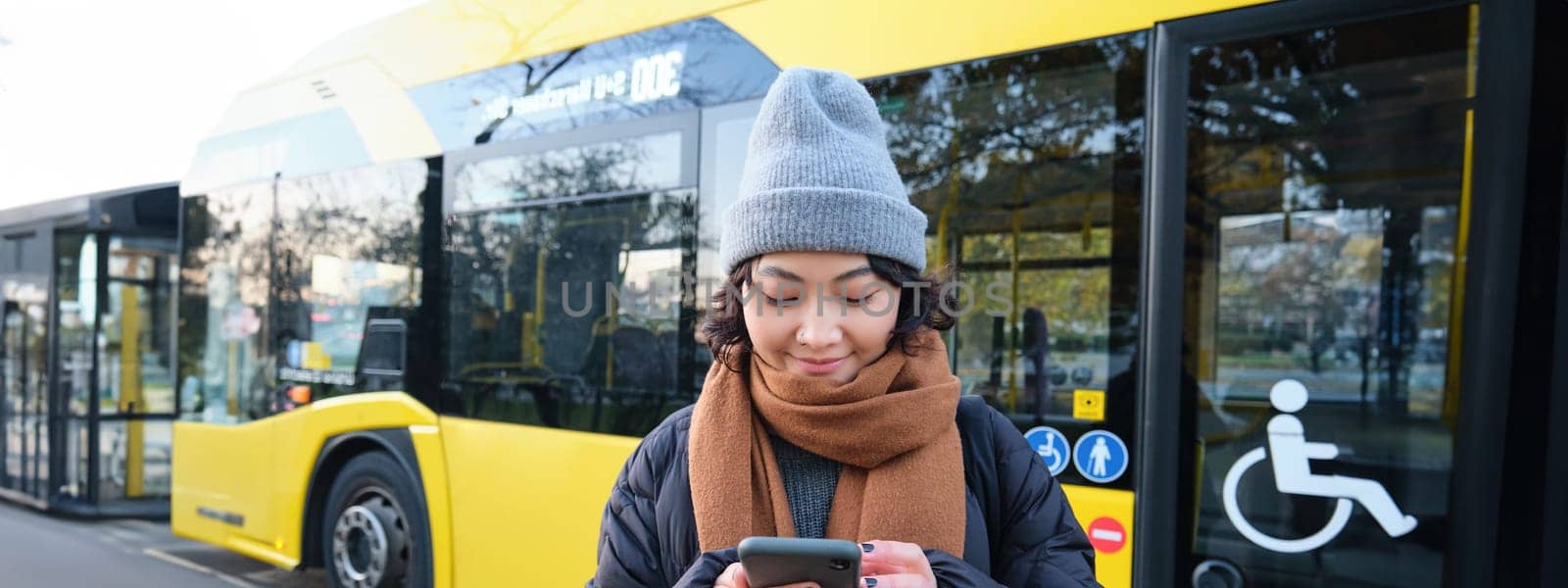 Image of girl student waiting for public transport, checks schedule on smartphone app, stands near city bus. Urban lifestyle concept