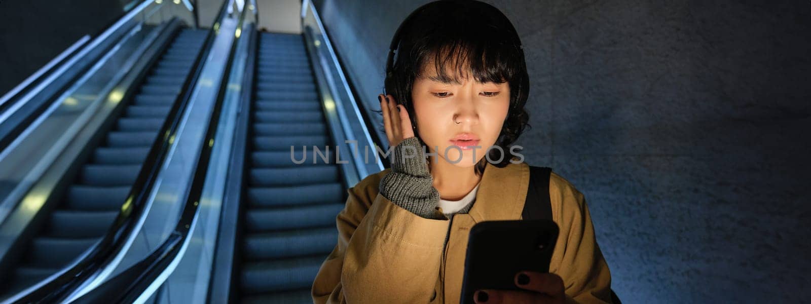 Woman in headphones, listens music, looks concerned at her phone screen, goes down escalator in city.