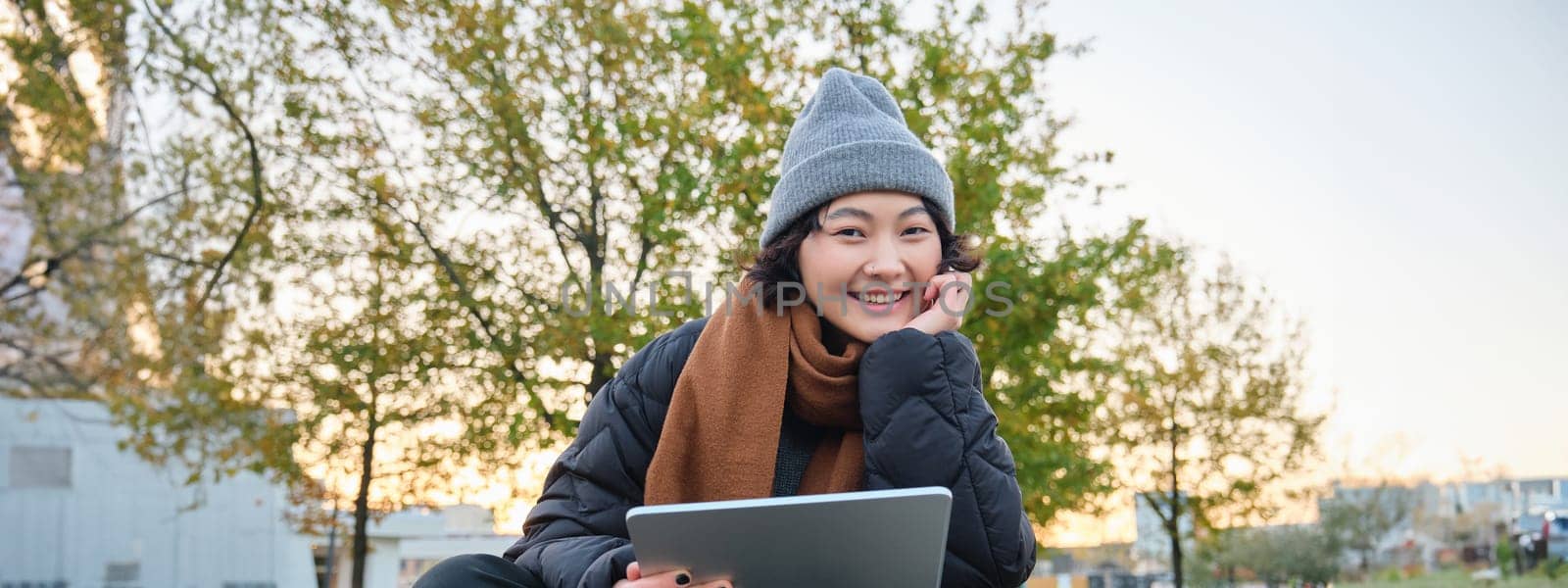Portrait of asian girl relaxing in park, watching videos or reading on digital tablet, sitting on bench in hat and scarf on chilly day, enjoing outdoors by Benzoix