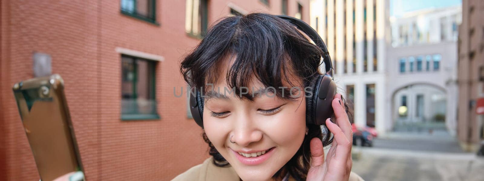Close up portrait of stylish brunette asian girl, listens music in headphones, touches earphones and smiles, enjoys favorite song, stands on street.