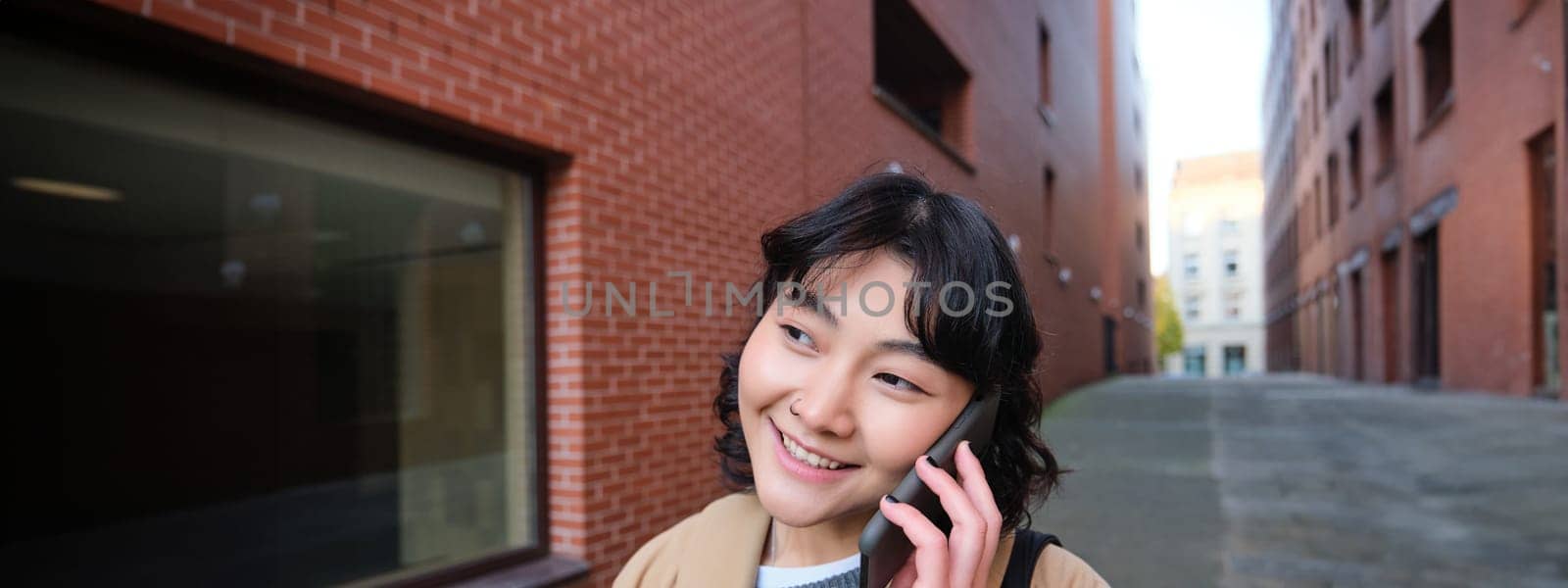 Cellular technology concept. Young modern girl talks on mobile phone, walks on street and smiles, has conversation over telephone by Benzoix