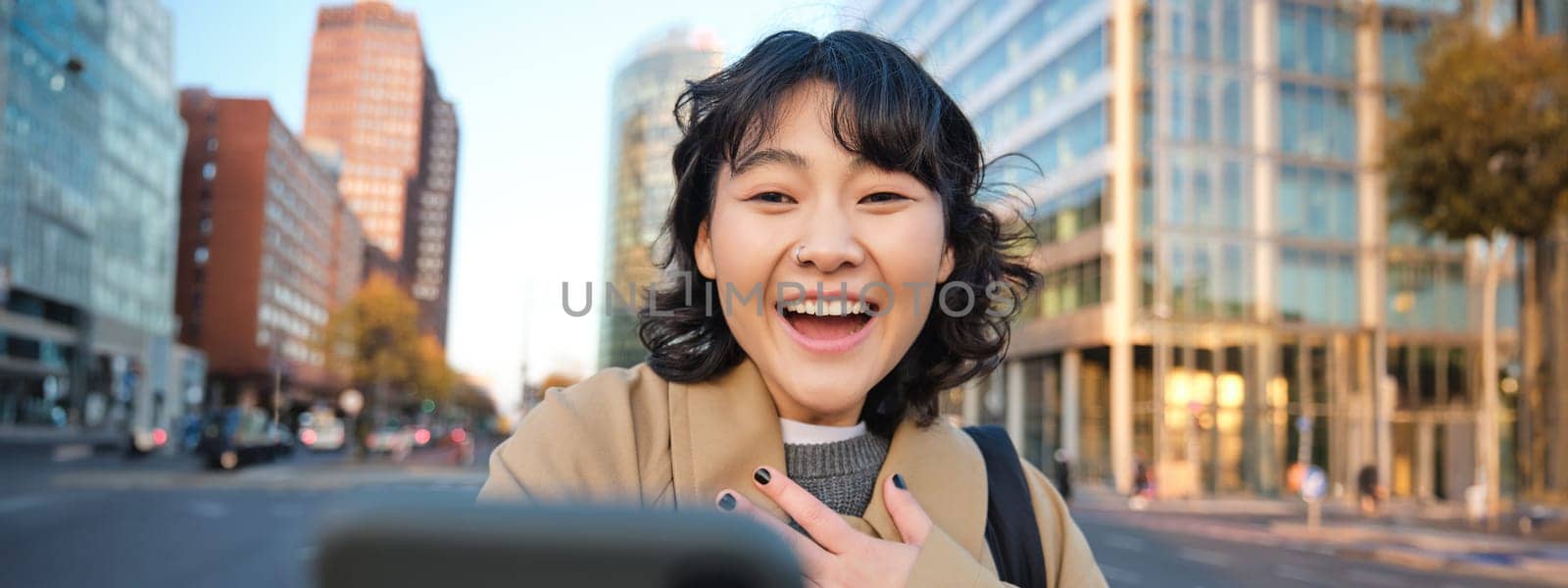 Image of korean girl video chats with smartphone, looks at her phone with surprised and amazed face, hears great news, stands on street.