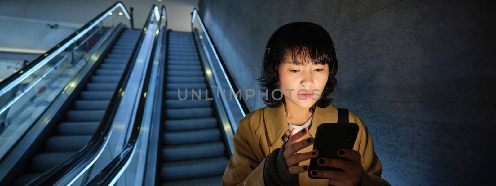 Urban lifestyle and people. Woman in headphones, looks serious at her smartphone app, checking map, public transport schedule with worried face, standing on escalator.