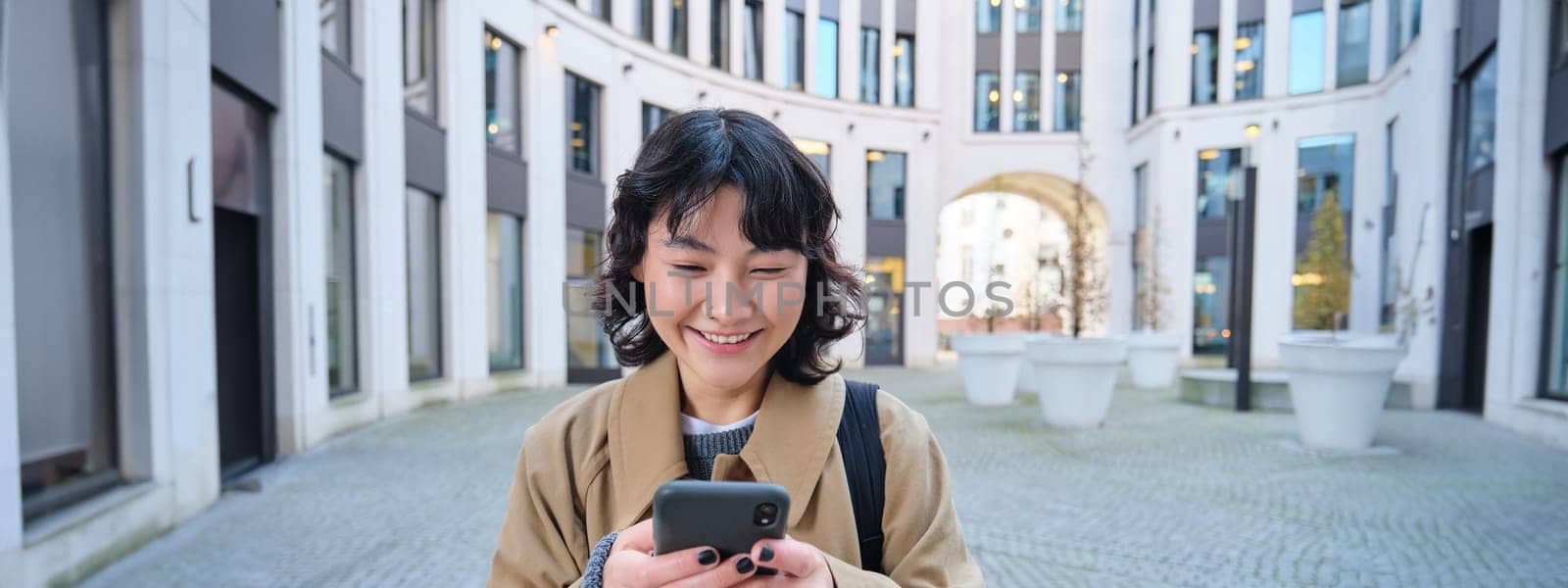 Stylish korean girl in headphones, listens music and uses mobile phone, stands in city centre, waits for someone on street and writes text message on her smartphone.