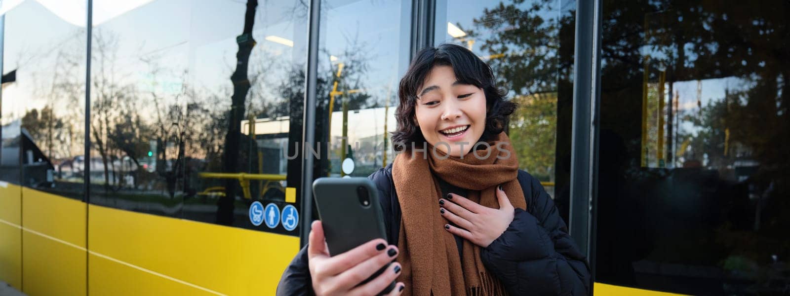 Portrait of cheerful asian girl talks on mobile phone, video chats, looks amazed at smartphone camera, stands on bus stop by Benzoix