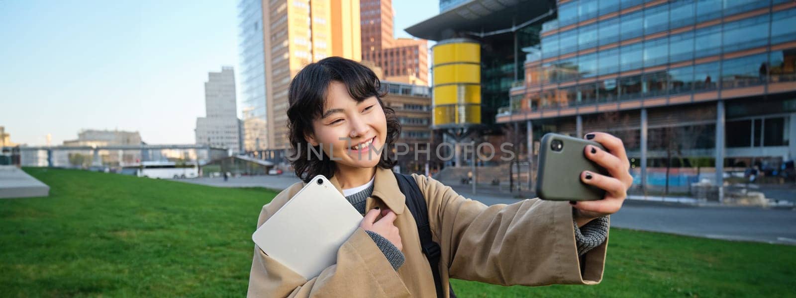 Happy university student, korean girl takes selfie with her papers and digital tablet, holds smartphone and poses near university campus by Benzoix