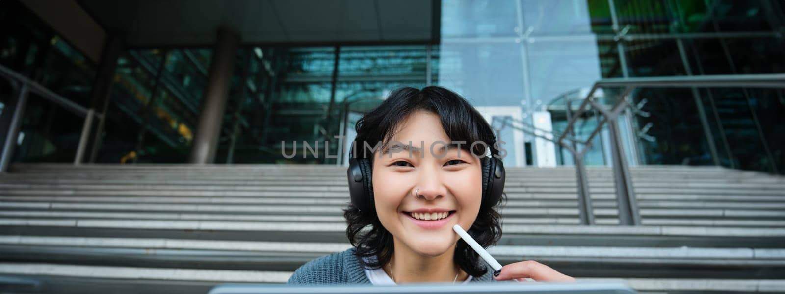 Close up of smiling asian girl feels inspired while draws on digital tablet, makes graphic design project, pencils user interface assignment, sits on stairs near campus.