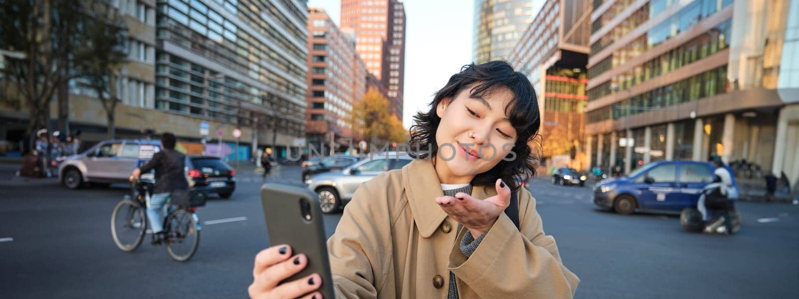 Cute korean girl, tourist takes selfie, video chats with friend and sends kisses to camera, records vlog on streets of city with smartphone by Benzoix