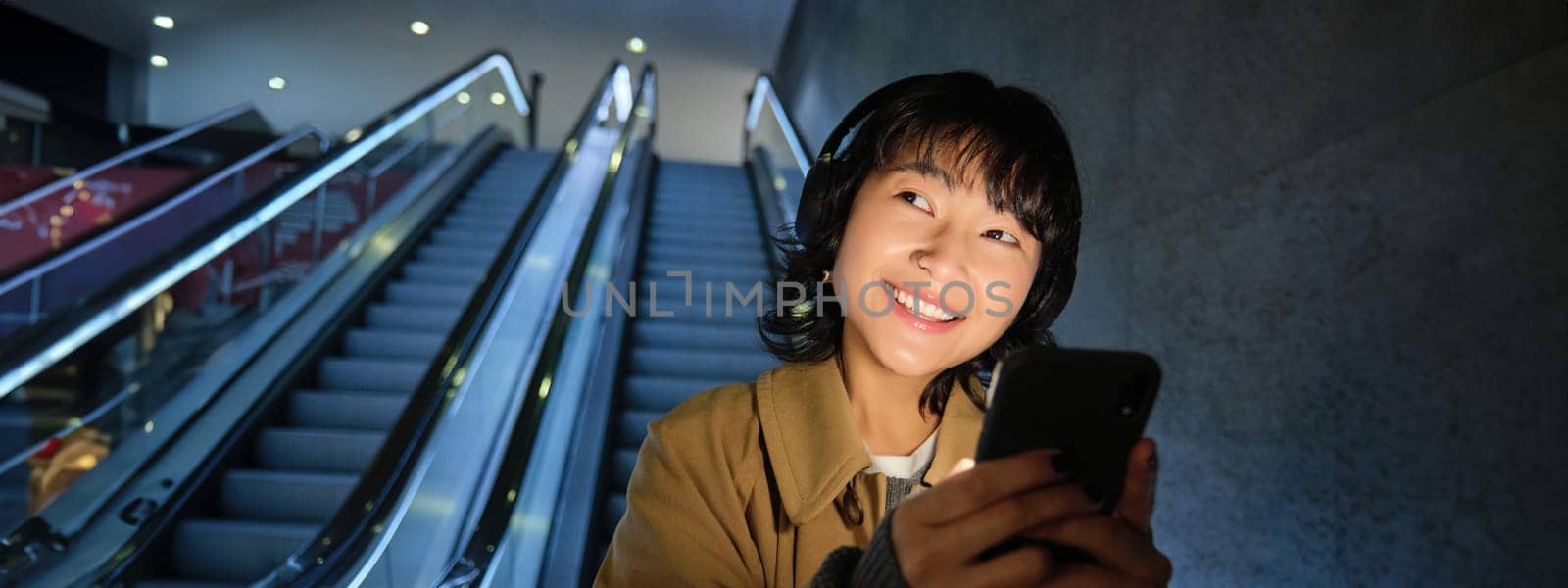 Smiling korean girl going down escalator in dark, holding mobile phone, using smartphone app, listening music, commuting in city.