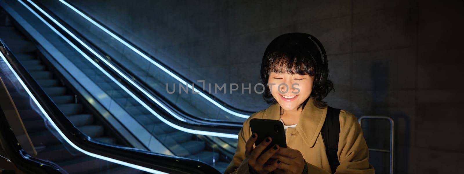 Happy asian woman in subway, standing near escalator, holding smartphone and listening music in headphones, smiling pleased by Benzoix