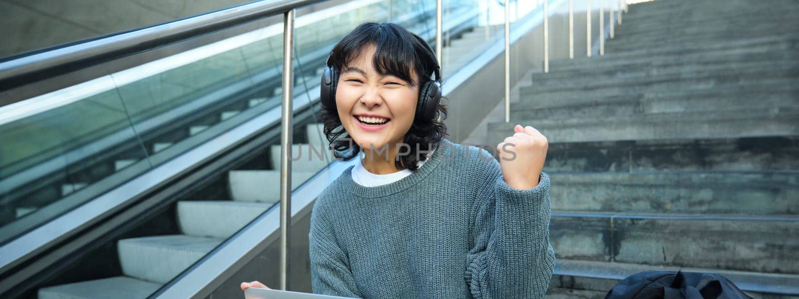 Cheerful korean girl in headphones, sits on stairs with laptop, celebrates, say yes, triumphs, receives good news, success on working project, works remotely on street by Benzoix