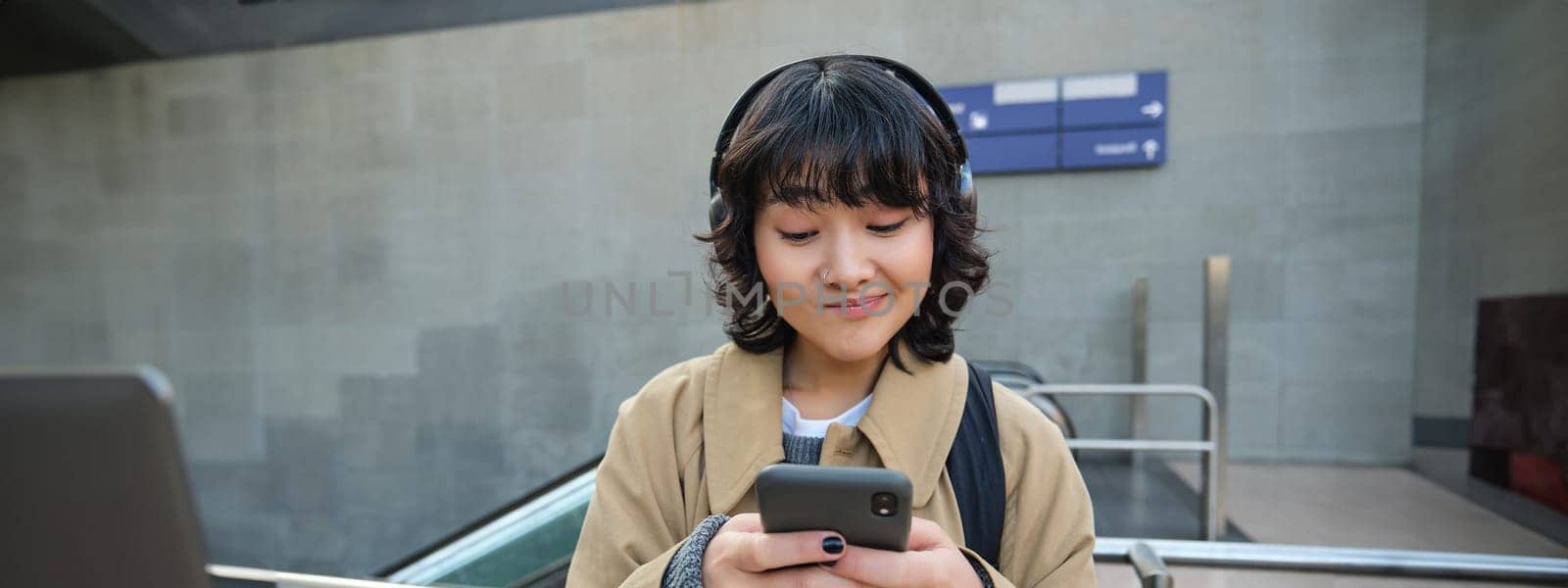 Technology and people. Young asian girl, student standing on street in headphones, listens music and smiling, chats on mobile phone, sends a text message, waits for someone on street.