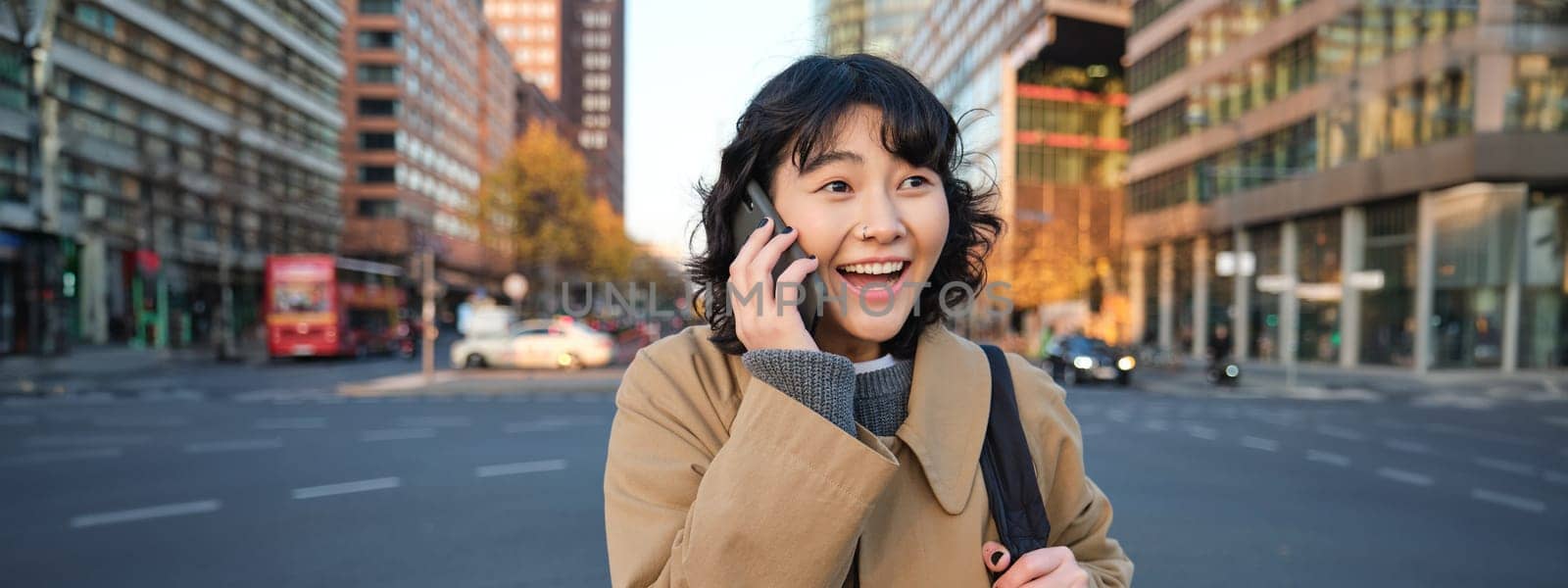 Portrait of happy korean girl, talks on mobile phone, looks surprised and happy, receives positive great news over telephone conversation, stands on street of city.