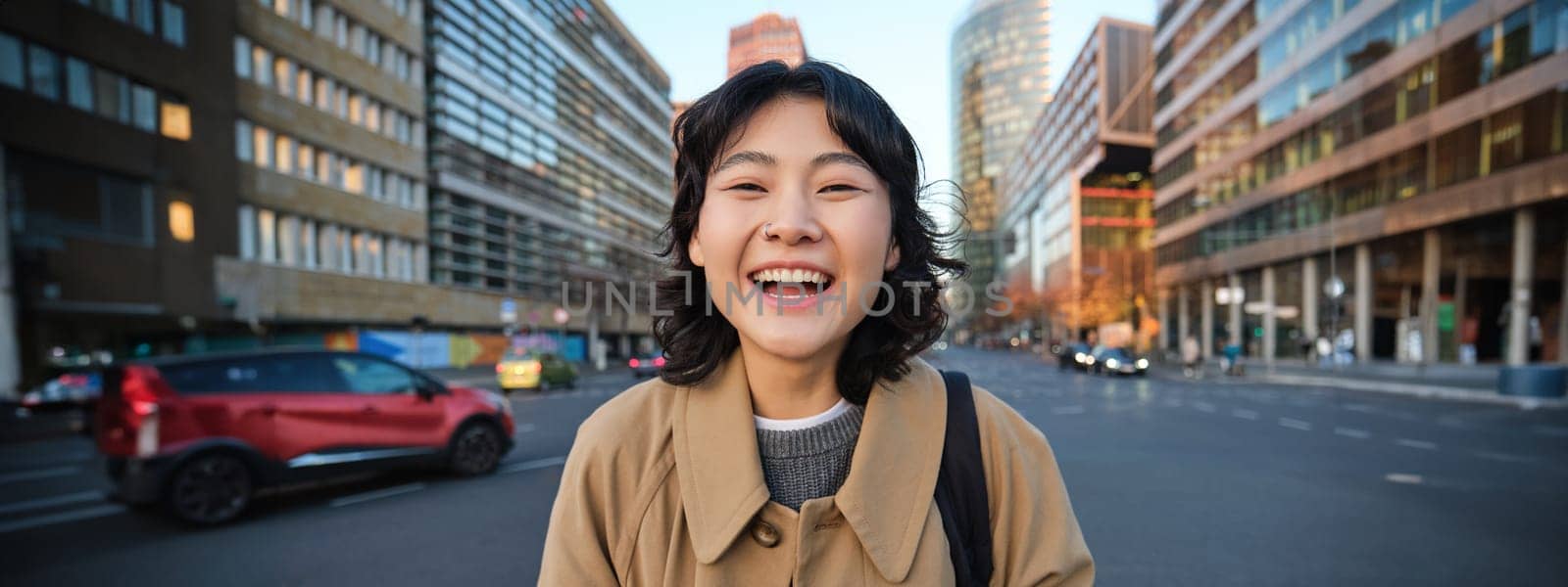 Portrait of asian girl student, stands in city centre with cars on busy street, holds digital tablet and smiles at camera by Benzoix