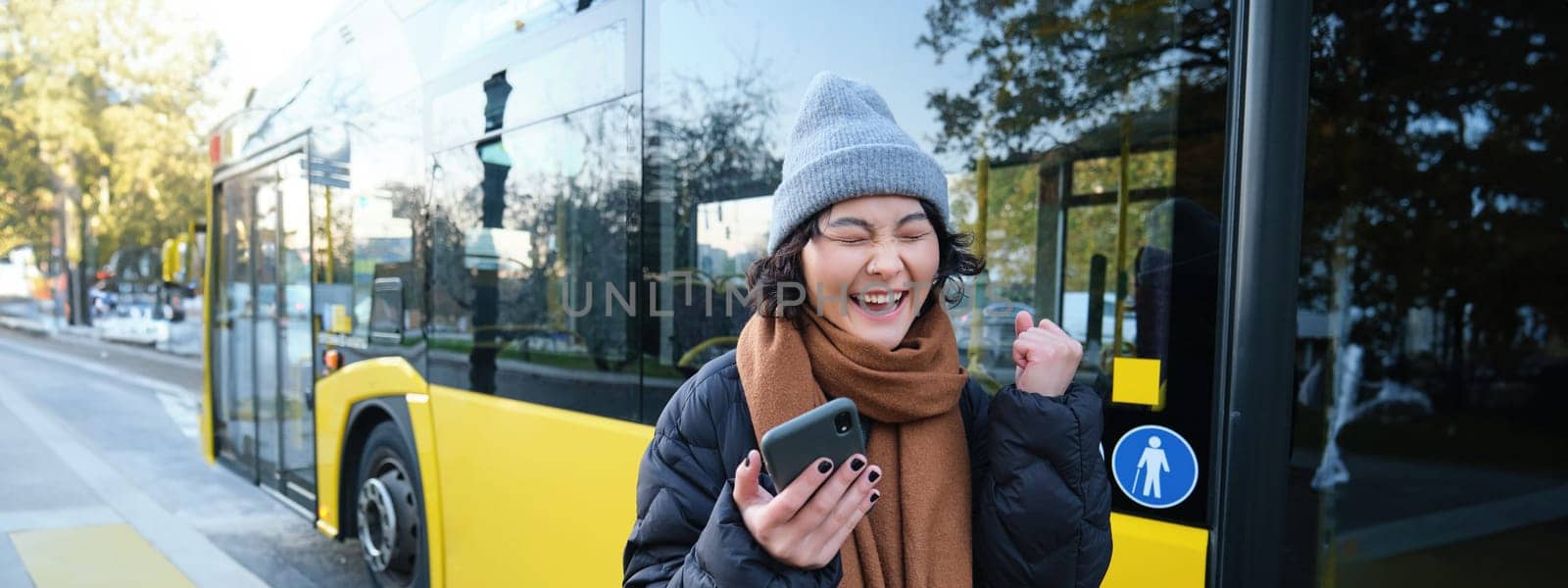 Modern people and lifestyle. Happy asian girl screams from joy, celebrates, stands near bus public transport and looks amazed.