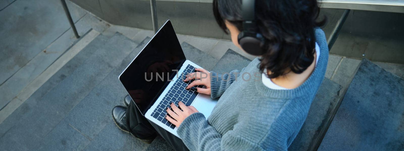 Close up portrait of girl, student works on laptop and listens music in headphones. Blank computer screen and hands typing on keyboard by Benzoix